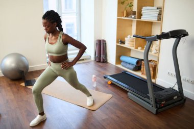 Afrika kökenli Amerikalı bir kadın, çeşitli spor aletleri ve ekipmanlarıyla iyi donanımlı ev spor salonunda paspasa atlıyor.