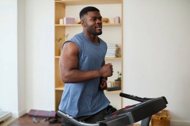 Modern bir odada koşu bandında koşan genç Afrikalı bir adam. Etrafı kitaplık ve kapalı bitkilerle çevrili. Egzersiz yapıyor.