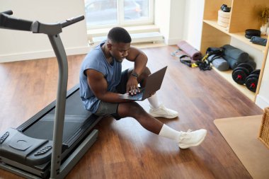 Spor salonunda dizüstü bilgisayarla çalışırken koşu bandına yaslanan Afrikalı Amerikalı bir adam. Çevreleyen çalışma ekipmanları organize bir fitness ortamı yaratıyor