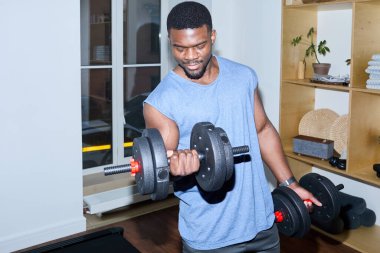 Kolsuz gömlekli Afro-Amerikan adam modern spor salonunda dambıl kıvırıyor.