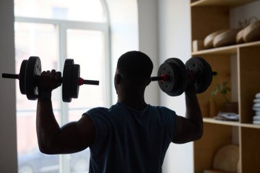 Spor salonundaki dambılları kaldıran adam büyük pencerenin yanında, fitness ekipmanları arka planda görünür.