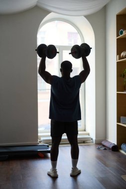 Parlak odada duran adam, dambılları azimle kaldırıyor ve spor yapmaya odaklanıyor.