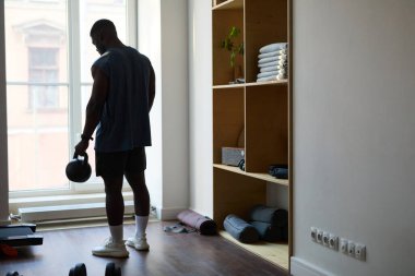 Ev jimnastik salonunda Kettlebell 'le çalışan bir adam. Oda geniş bir pencere, spor aletleriyle ahşap raflar ve ahşap zemin içeriyor.