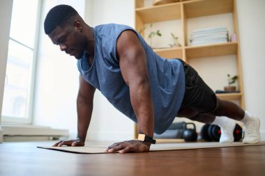 Afrika kökenli Amerikalı bir adam spor kıyafetleri içinde egzersiz paspası üzerinde şınav çekiyor ve spor yapmaya konsantre oluyor.