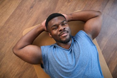 Egzersiz yaptıktan sonra yoga minderinde uzanan Afrikalı Amerikalı adam, kollarını başının arkasında dinlendiriyor ve rahatlamış görünüyor.