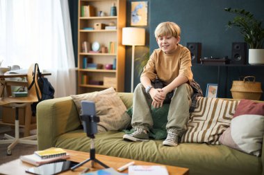 İyi dekore edilmiş bir oturma odasındaki kanepede oturan genç çocuk, rahat ve mutlu görünüyor. Kitaplar, bitkiler ve aletler rahat bir atmosfer yaratır.