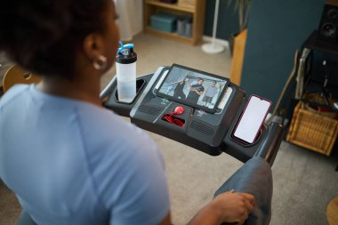 Koşu bandında spor yapan bir kişi egzersiz videosu gösteren bir tabletle spor yapıyor, spor ekipmanlarıyla çevrili ve kapalı bir ortamda ekrana odaklanıyor.