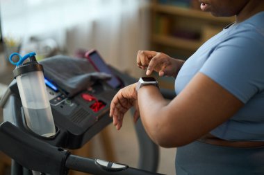 Genç bir kadın koşu bandında su şişesi ve yakınlarda akıllı gözlerle antrenman yapıyor. Çevrede çeşitli kişisel eşyalar ile kapalı ortamdaki fitness hedeflerine odaklanmış