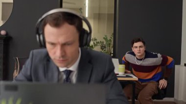 Genç beyaz erkek çalışanın ofis bilgisayarında bilgisayarla kulaklıklı yetişkin meslektaşına yardım ederken orta boy fotoğrafı.
