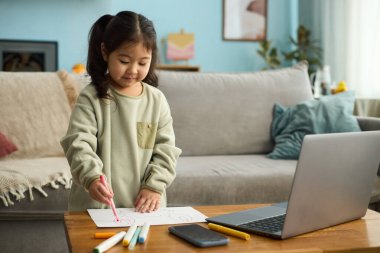 Masada keçeli kalemlerle oturan genç bir kız gülümserken, dizüstü bilgisayar ve akıllı telefon ahşap masanın üzerinde, arka planda kanepede yatarken.