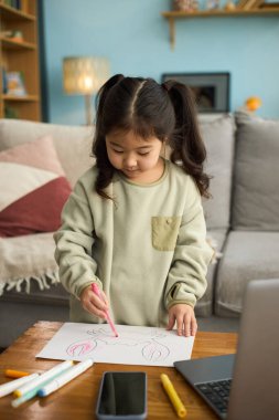 Beyaz kağıda renkli kalemler çizen Asyalı genç bir kızın portresi. Oturma odasındaki ahşap masada dururken.