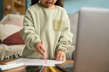 Masanın yanında duran pembe kalemle çizim yapan çocuk, rahat ev ortamında heyecan ve yaratıcılık gösteriyor
