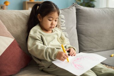 Kanepede oturan küçük kız beyaz kağıda sarı boya kalemi çizmeye odaklanmış, konsantrasyon ve özenle yaratıyor.