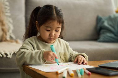 Renkli kalemlerle çizime dalmış çocuk, rahat oturma odasında masada oturuyor, sakin ve rahat bir ortamda yaratıcı sanat eserleri yaratıyor.