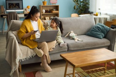 Anne telefonunu kontrol ederken, kız yanında oturuyor, iletişim kuruyor, kitaplarla, bitkilerle ve oyuncaklarla sıcak bir ortam yaratıyor.