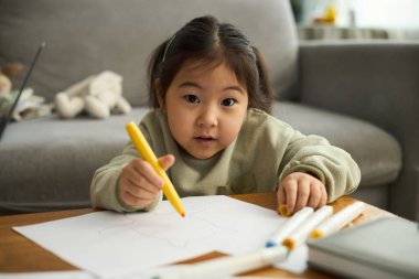 Genç Asyalı bir kızın portresi. Aktivite çiziyor, elinde odaklanmış bir ifadeyle sarı bir boya kalemi tutuyor.