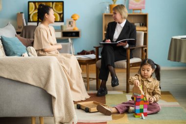 Çocuk zemindeki yapı taşlarıyla oynarken terapist ve kadın müşterilerle danışmanlık oturumu gerçekleşiyor.