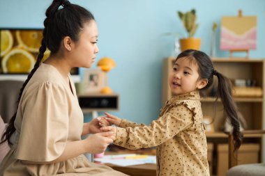 Uzun örgülü bir kadın oturma odasında genç bir kızla el ele tutuşuyor. Duvarlarda sıcak dekor ve sanat eserleri var. İkisi de birbirine bakıyor.