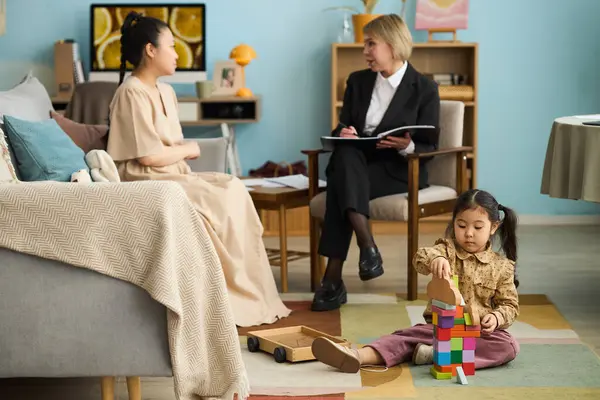 Çocuk zemindeki yapı taşlarıyla oynarken terapist ve kadın müşterilerle danışmanlık oturumu gerçekleşiyor.