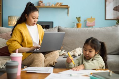 Genç kızı yakınlardaki masada çizim yaparken Asyalı kadın dizüstü bilgisayara odaklanıyor.