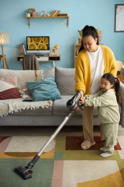 Sarı hırkalı anne ve yeşil eşofmanlı kız modern dekorlar, raflar ve sanat eserleriyle renkli oturma odalarını süpürüyorlar.