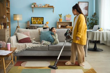 Bir kadın oturma odasını süpürürken çocuk kanepenin, modern dekorun, ahşap mobilyaların ve geniş pencerelerin keyfini çıkarıyor.