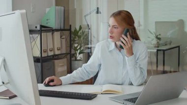 Beyaz kadın çalışanın ofis masasında bilgisayar kullanırken cep telefonu kullanırken orta boy fotoğrafı.