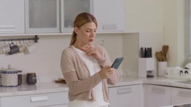 Orta boy beyaz bir kadının ev mutfağında cep telefonuyla sohbet ederken görüntüsü.