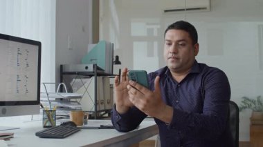 Yetişkin Asyalı erkek müdürün ofisinde otururken akıllı telefonunu kontrol ederken orta ölçekli bir fotoğrafı.