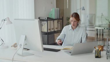 Ofiste dizüstü bilgisayarla çalışırken beyaz elbiseli genç bir iş kadınının monitör ekranının önünde oturduğu orta ölçekli bir fotoğraf.