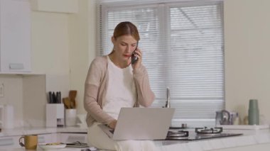 Kadın beyaz girişimcinin ev mutfağında dizüstü bilgisayarla konuşurken cep telefonuyla konuşması.