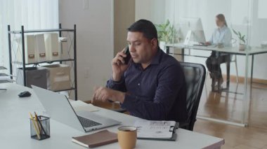 Ofisteki kablosuz bilgisayarda çalışırken Asyalı erkek uzmanın akıllı telefondan konuştuğu orta yavaş bir çekim.