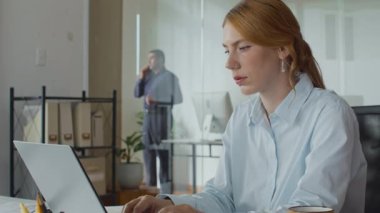 Beyaz kadın çalışanın kablosuz bilgisayarla çalışırken, erkek meslektaşın ofiste telefon görüşmelerini çekerken.