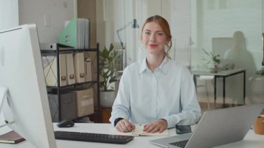 Resmi giyinmiş neşeli beyaz kadın girişimcinin ofis masasında otururken kameraya gülümseyerek bakışının orta boy portresi.