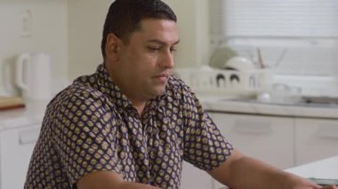 Orta yaşlı Asyalı bir adamın rahat ev mutfağında kablosuz bilgisayarla çalışırken çekilen görüntüsü.