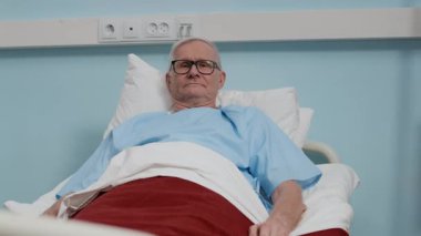 Hastane yatağında dinlenirken kameraya bakan, gözlüklü, yaşlı, beyaz bir adamın orta boy fotokopi portresi.