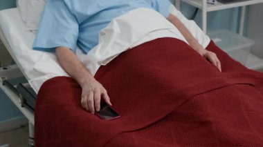 Akıllı telefonlu yaşlı beyaz adamın portresini yukarı kaldır ve klinik koğuşundaki kameraya bak.