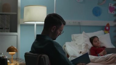 Hasta kızın hastane koğuşunda dijital tablet kullanarak otururken kitapla uyuyakalan yorgun beyaz bir baba fotoğrafı.