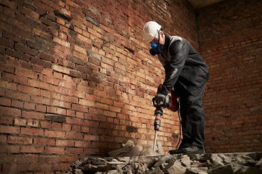 Kirli üniformalı beyaz adamın düşük açılı görüntüsü. Kasketli ve solunum cihazlı. Beton zemini matkapla tahrip ediyor.