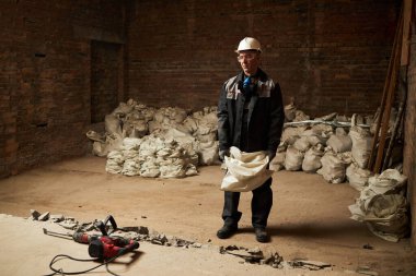 Kirli üniformalı, yorgun, beyaz bir adam elinde ağır bir çimento torbasıyla odanın ortasında kırık zemin ve çimento torbalarıyla dikiliyor.