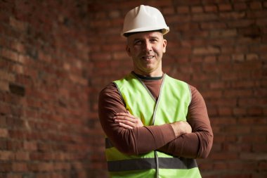 Kameraya gülümseyen beyaz ustabaşının portresi. Arka planda tuğla duvarlar olan odada çapraz kollarla fotoğraf çektirirken.