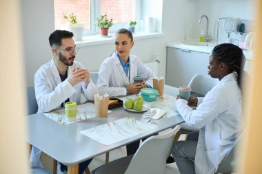 Üç tıp uzmanından oluşan bir grup, tatilde hastane kafeteryasında oturmuş laboratuvar önlüğü giyip kahve fincanları tutarak fikirleri tartışıyor.