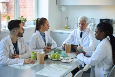 Farklı geçmişlere sahip doktorlar öğle yemeği odasında tıbbi bulguları tartışıyor. Sağlıklı yemek seçenekleri ve kahve fincanları da dahil.