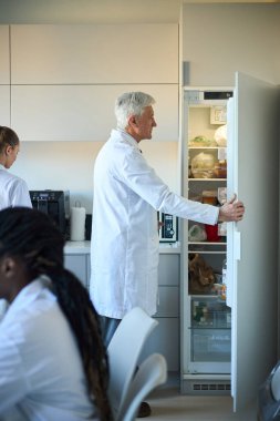Meslektaşlar çalışırken buzdolabını açan yaşlı bir bilim adamı. Laboratuvar ekipmanları ve malzemeleri görünür, etkin araştırma ortamı
