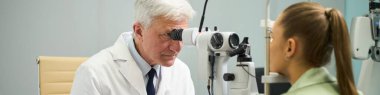 Laboratuvar önlüğü giyen yaşlı erkek doktor tıp bürosunda uzman göz muayenesi cihazı kullanarak genç kadınların gözlerini inceliyor.