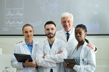 Çeşitli tıp uzmanları laboratuvar önlükleriyle birlikte duruyorlar, bazıları tabletleri tutuyor ve gülümsüyor, arkadaşlığı ve takım çalışmasını tıbbi bir ortamda canlandırıyorlar.