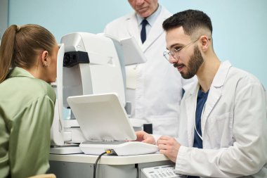 Göz muayenesine giren bir kadın bir göz doktoruyla klinik ortamda tıbbi ekipman kullanıyor. Kıdemli doktor prosedürü arka planda izliyor.