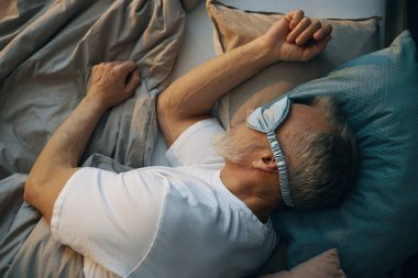 Yaşlı, beyaz bir adam, uyku maskesiyle uyuyarak gözlerini kapatıyor, huzur içinde yatarken yanında yastıklar ve onu çevreleyen yataklarla yatıyor.