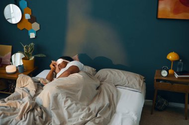 Sıcak yatak odasında, modern dekorlar ve duvarlarda parlayan ılık ışıklarla uyandıktan sonra yatakta uzanan bir adam, sabah keyfine bakarken sakin bir atmosfer yaratıyor.