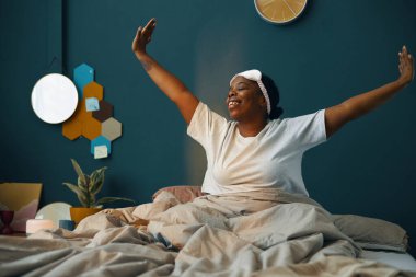 Sıcak yatak odasında sabah esneme hareketleri yaparken gülümseyen bir kadın. Etrafı sıcak dekor ve altıgen duvar resimleriyle çevrili yeşil duvarlarla çevrili. Rahatlatıcı bir atmosfer yaratıyor.